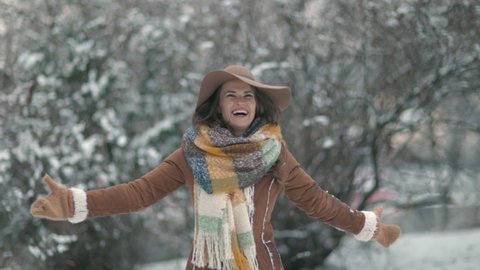 happy stylish woman in brown hat and scarf with mittens in sheepskin coat rejoicing outside in the city park in winter.