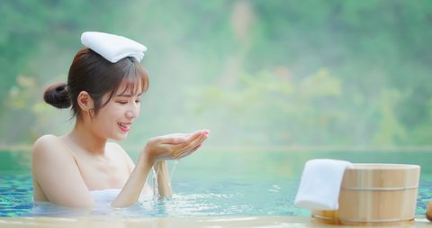 Slow motion of asian woman scoop up the water and feel the drop while sitting in hot spring with towel on head