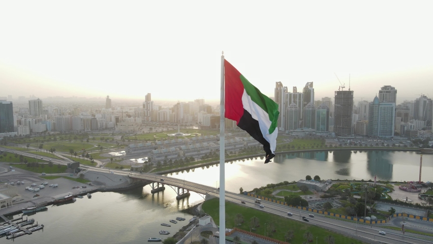 The Flag of the United Arab Emirates waving in the air, the Blue sky and city development in the Background, The national symbol of UAE over Sharjah's Flag Island. 4k Footage