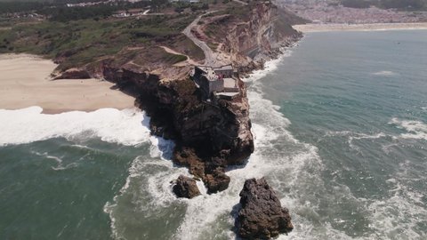Waves crashing on rocky cliffs with Fortress of Saint Michael the Archangel and Nazare lighthouse suited on top.