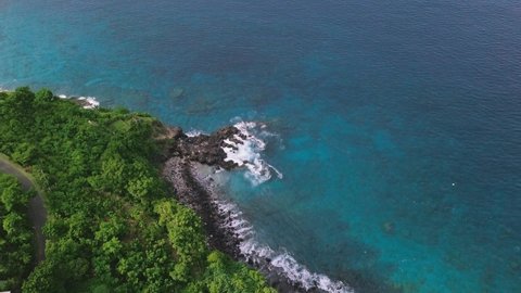 Aerial view of coastal cliffside with blue transparent water showing the corals undersea. Full of greenery, emerald blue ocean, waves crashing to the rocky shoreline and vast of corals underwater.