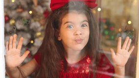 portrait of a little cute girl in a red dress with a bow on her head through glass close up. Merry Christmas and Happy Holidays. Happy New Year. Hygge. Winter. High quality 4k footage
