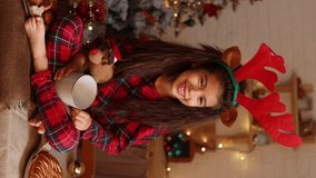 little brunette girl in checkered red pajamas with reindeer horns on her head is eating a Christmas cake and drinking tea from a white mug in a beautifully decorated kitchen. High quality 4k footage