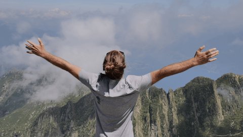 Young man stands on mountain top above clouds, arms wide open. Young hiker reaches the top, celebrating with his arms raised. Achievements concept 