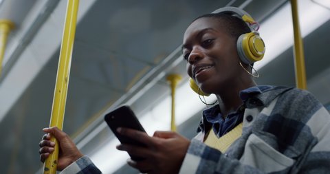 Cinematic close up of young smiling african woman listening to the music with smartphone while traveling by train in subway. Concept of transportation, technology, connection, communication, lifestyle