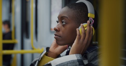 Cinematic shot of young african woman with headphones relaxing by listening to music while traveling by train in subway. Concept of entertainment, transportation, technology, tourism, inspiration.