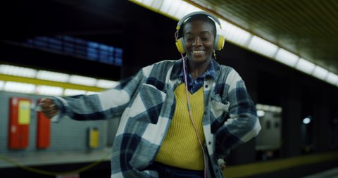 Cinematic shot of young african woman with headphones listening to music and having fun to dance alone while waiting for train at subway station.Concept: transportation, technology, lifestyle, fashion