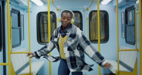Cinematic shot of young african woman with headphones listening to music and having fun to dance alone while traveling by train in subway. Concept of transportation, technology, lifestyle, fashion.