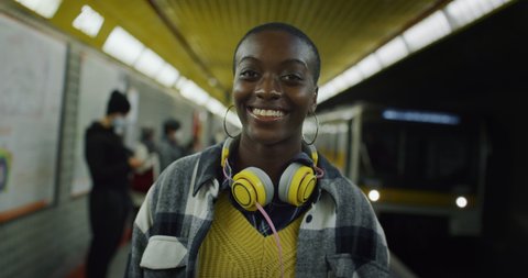 Cinematic shot of young african woman with headphones is smiling in camera while waiting for train at subway station. Concept of transportation, technology, lifestyle, fashion, urban, monopolies.
