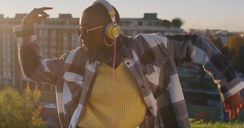 Cinematic shot of young stylish african woman with yellow headphones listening to music and having fun to dance alone on urban scape background at sunset.