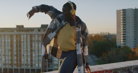 Cinematic shot of young stylish african woman with yellow headphones listening to music and having fun to dance alone on urban scape background at sunset.
