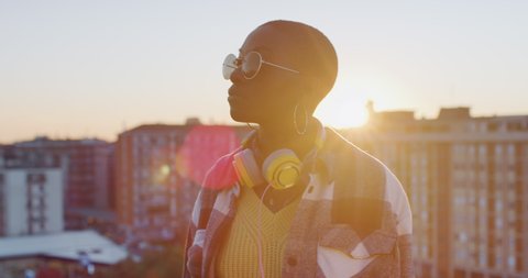 Cinematic shot of young happy stylish african woman with sunglasses and headphones is feeling free and enjoying view on urban scape background at sunset.