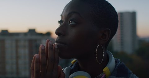 Cinematic close up shot of young serene peaceful african woman keeping hands together while meditating and feeling free on urban scape background at sunset.