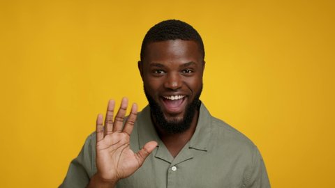 Hi. Portrait Of Positive Cheerful Black Guy Waving Hand At Camera, Friendly Young African American Man Greeting Somebody And Saying Hello While Standing Over Yellow Studio Background, Slow Motion