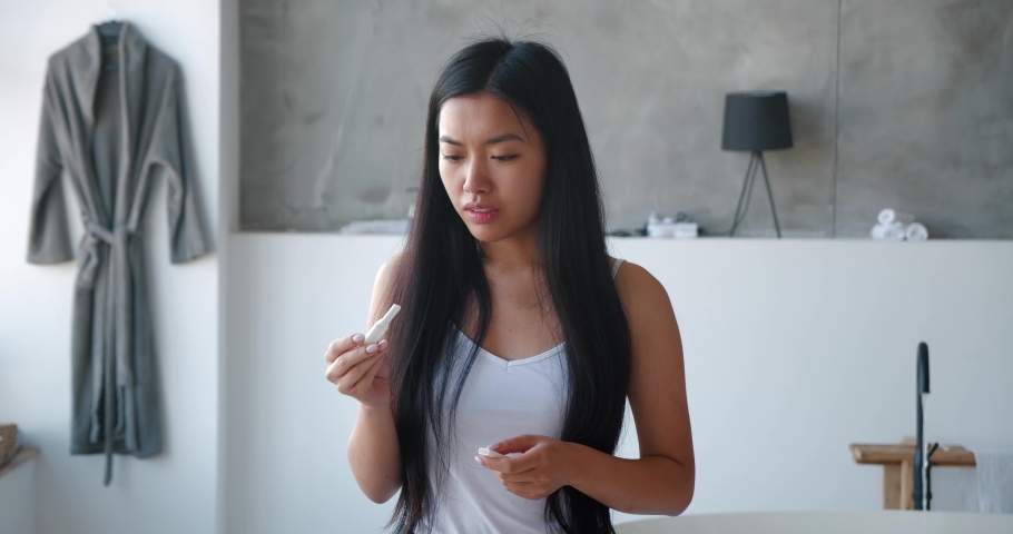 Young Asian woman holding pregnancy test strip waiting for results while standing in bathroom feeling worried and frustrated after results. Contraception, unwanted pregnancy, infertility concept | Shutterstock HD Video #1082037941