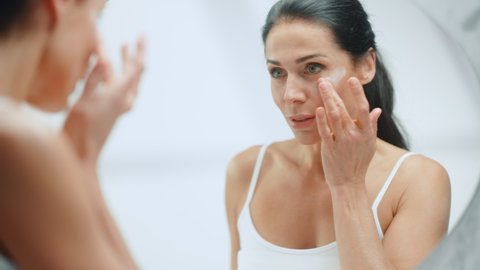 Portrait of Beautiful Caucasian Woman Gently Applying Face Cream Mask with Sensual Gestures, Looking in Bathroom Mirror. Middle Aged Female Makes Skin Soft with Natural Cosmetics Skincare Product