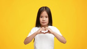 Cute Little Korean Girl Showing Heart Symbol With Fingers Posing Standing Over Yellow Background, Smiling To Camera. Studio Shot Of Asian Kid Gesturing Heart Shape Symbolizing Love. Slowmo