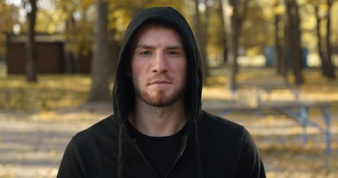Young caucasian man wearing black hoodie raising head and looking seriously to camera, posing outdoors in autumn public park, slow motion