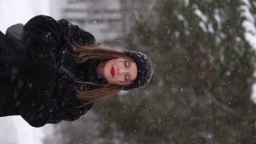 Vertical video of beautiful happy attractive european woman enjoying winter snowy weather outdoors in cold white frosty landscape. Video portrait of happy woman walking happily under falling snow