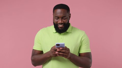 Dreamful happy young bearded african american man 20s wears green t-shirt hold using mobile cell phone typing browsing chatting send sms isolated on plain pastel light pink background studio portrait