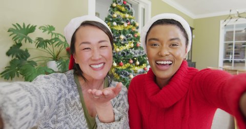 Happy diverse female friends in santa hats making christmas video call, blowing kisses and smiling. christmas, festivity and communication technology.