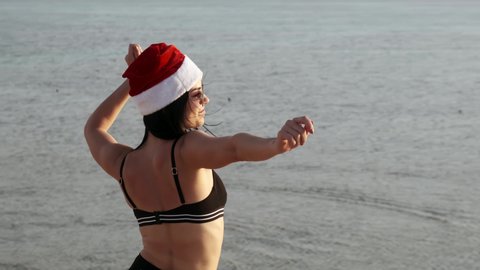 Young brunette woman in red santa hat with long hair resting on the sea, the lady is happy Christmas new year and tries to rush to look at the seascape
