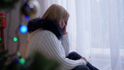 Side view depressed lonely woman sitting on Christmas alone at New Year tree in living room crying. Sad frustrated Caucasian adult lady indoors at home on holiday. Loneliness and holidays concept