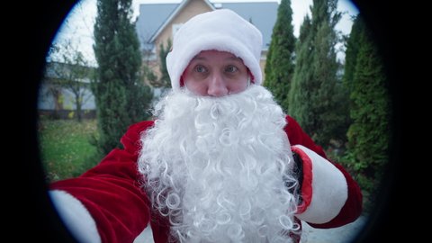 View through peephole of Santa Clause ringing doorbell standing on green front yard outdoors. Front view man in red Father Christmas costume with white beard and gift bag coming on New Year