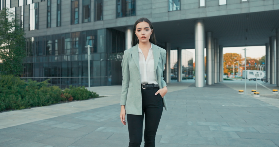 Attractive girl with brown hair walks out of corporate building for break, dressed in smart shirt and jacket imposed on back, walks outside past glass skyscraper, secretary, businesswoman, boss