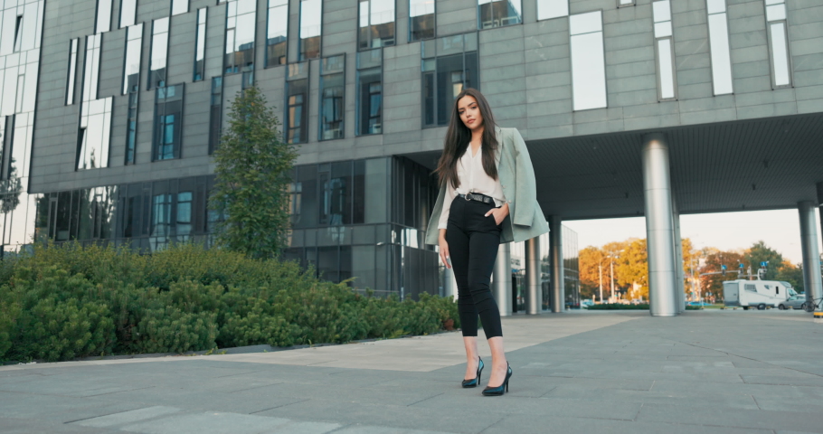 Beautiful woman with magnificent figure stands in front of modern office building dressed in elegant clothes stilettos, businesswoman, head of company, crosses arms on chest serious face
