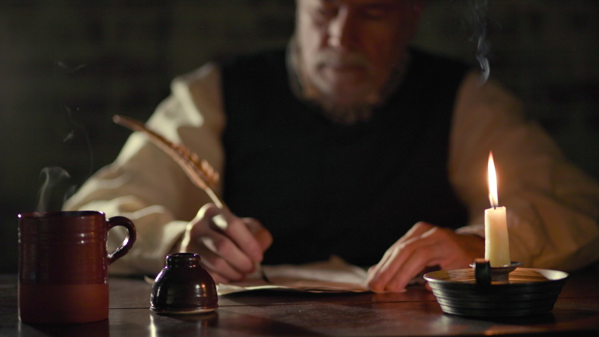 Scene of a mature man in the 18th century lit by candlelight sitting at a desk with a hot beverage writing with a quill pen. Royalty-Free Stock Footage #1082060588