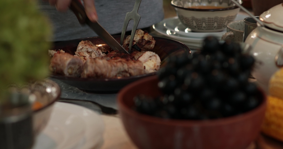 Hands cut stuffed turkey roll using knife and two prong fork on Thanksgiving dinner table with vintage crockery and autumn fruits. Traditional Thanksgiving turkey roulade roasted on bbq grill