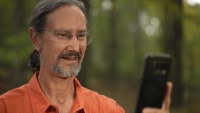 Portrait of ethnic, mature man smiling and talking to someone on video chat on smartphone in a natural outdoor environment.