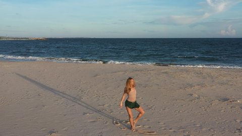 Young beautiful girl ballerina is dancing on the beach near the sea on the golden sunset. Moving woman, charming actress smile. Drone flies near dancer on the ocean shore. 