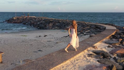 Young beautiful girl ballerina is dancing on the beach near the sea on the golden sunset. Moving woman, charming actress smile. ballerina dancing along the path by the sea, drone follow her