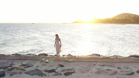 Young beautiful girl ballerina is dancing on the beach near the sea on the golden sunset. ballerina is dancing along the path by the sea, drone follows her. back sunset light