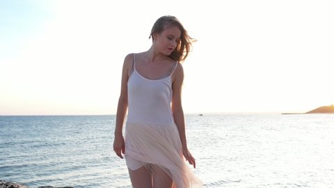 Young beautiful girl ballerina is dancing on the beach near the sea on the golden sunset. Moving woman, charming actress smile. Drone flies near dancer by ocean. ballerina dresses, straightens dress