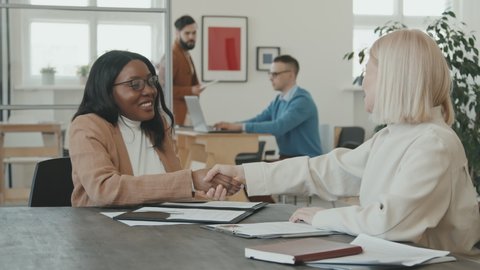 PAN shot of happy African-American female job candidate singing contract and shaking hands with female HR manager in office