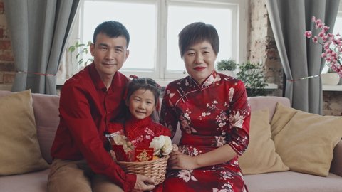 Portrait shot of happy Chinese family in traditional clothing sitting on couch at home and holding Lunar New Year gift basket while posing for camera
