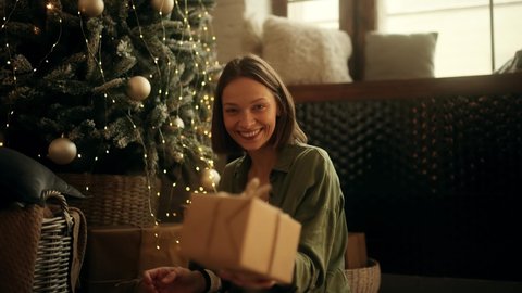 Happy attractive brunette woman holding and showing a gift to the camera. Caucasian beautiful woman sitting at the Christmas tree.