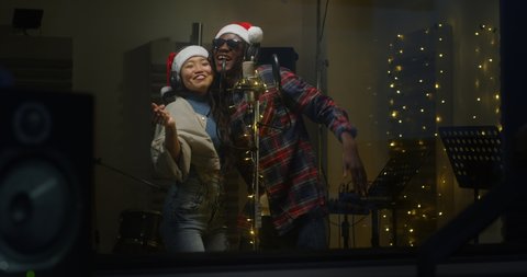 Cinematic shot of young professional smiling energetic band singers with santa hats are performing together a new christmas song with a microphone while recording it in a music studio with manager.