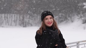 Video footage of beautiful happy woman enjoying winter snowy weather outdoors in cold white frosty landscape. Video portrait of happy woman blowing kiss into camera happily under falling snow