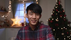handsome young asian man looking at lens talking on video chat online with christmas tree in background. guy face camera waving hands greeting family on internet virtual phone call