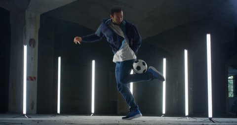 Cinematic shot of young arab freestyle soccer player is juggling sport ball with his knee and feet while training tricks of self-expression with football in underground parking with neon lights.