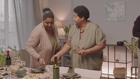 Medium long of two younger and older African American women putting cutlery down on dinner table for holiday celebration, cropped Black men drinking wine and talking on background