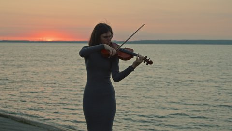 Young woman musician is playing violin outside, performing solo on rivershore, pink sunrise is on a background, autumn morning, Slow motion.
