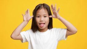 Misbehavior. Portrait Of Funny Little Korean Girl Sticking Out Tongue Holding Hands Near Temples Posing And Grimacing Over Yellow Background, Looking At Camera. Studio Shot