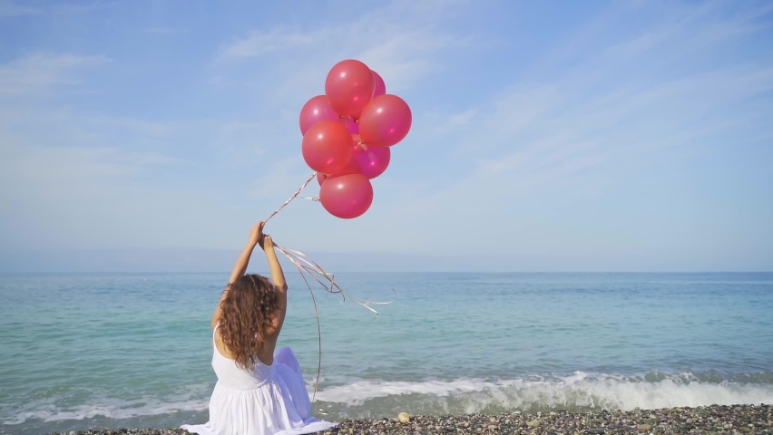 Happy Girl in white dress holds red balloons on the sea and the beach background. Young woman celebrates her birthday on the beach, rejoices, runs, smiles, laughs