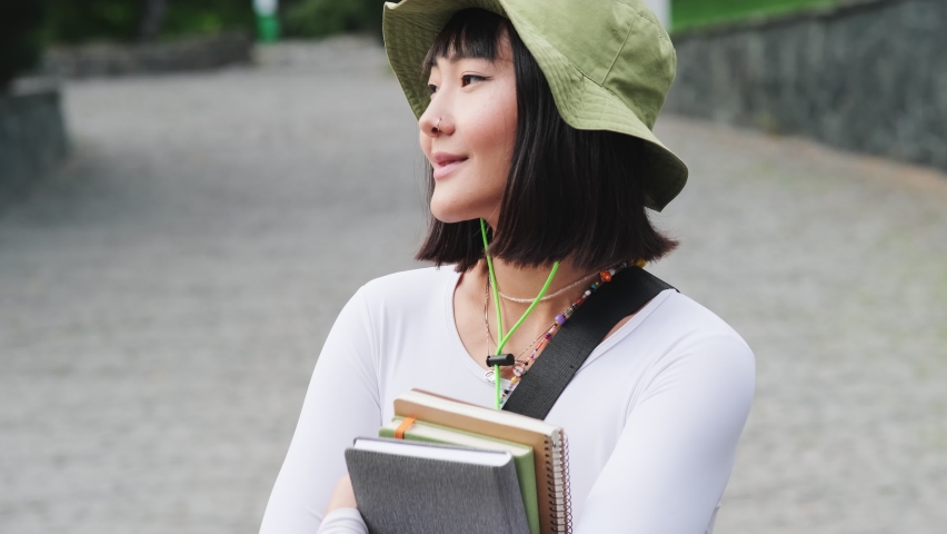 Smiling Asian woman in panama standing with a stack of textbooks on the street