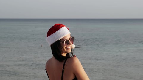 Young brunette woman in red santa hat with long hair resting on the sea, the lady is happy Christmas new year and tries to rush to look at the seascape